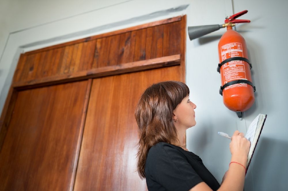Woman inspecting a wall-mounted fire extinguisher and recording notes during a fire risk assessment 